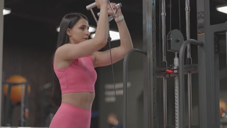 A-hispanic-brunette-woman-in-a-pink-suit-pulls-a-rope-in-a-crossover-with-her-hands-to-train-her-shoulders.-Shoulder-workout-in-a-trainer.-Professional-woman-instructor